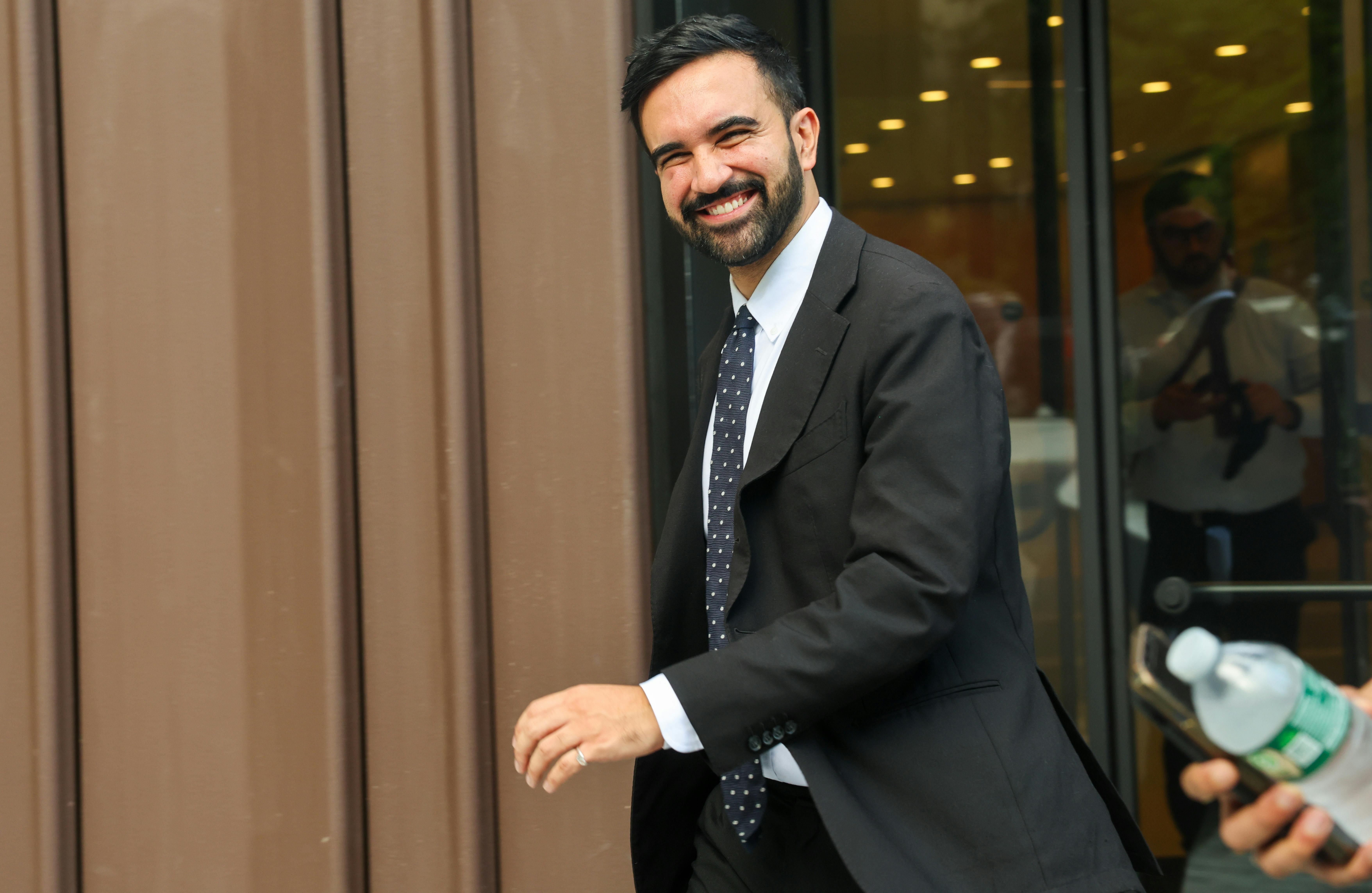 Zohran Mamdani smiles while walking out of a building