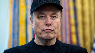 Elon Musk wears a black DOGE cap and purses his lips. He is sporting a black eye.