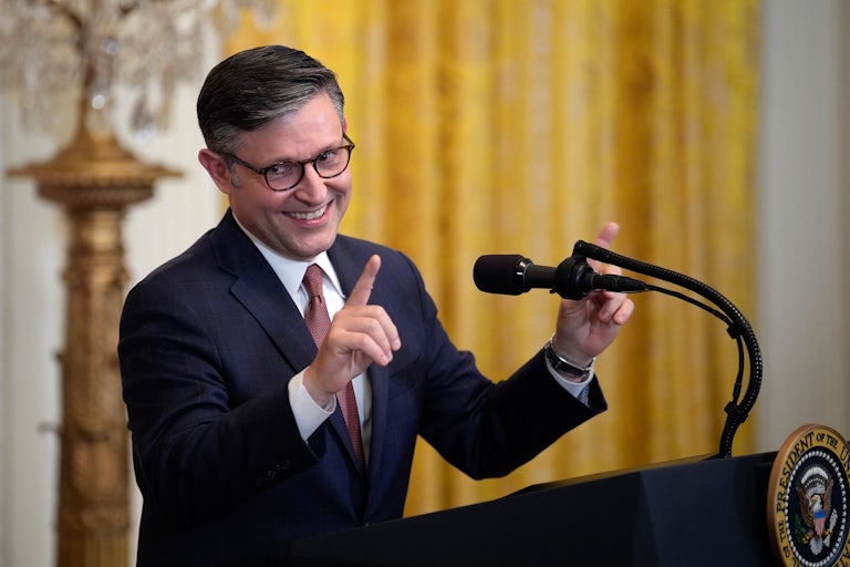 House Speaker Mike Johnson speaks at the presidential podium and smiles while pointing both index fingers.