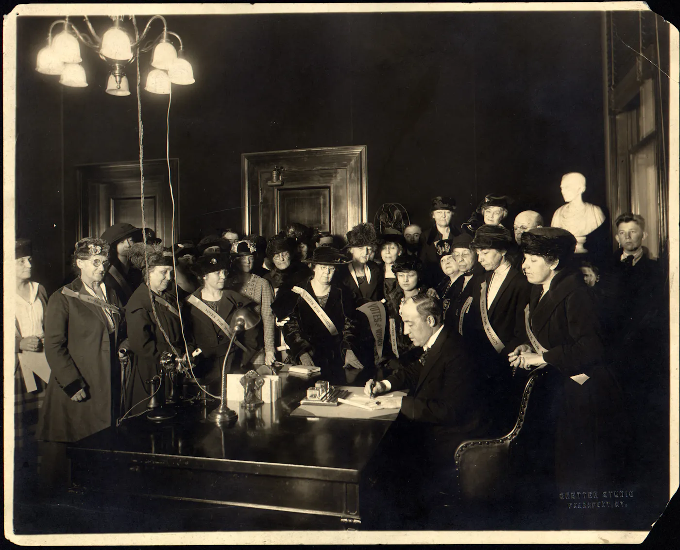 Photograph of suffragists in 'Votes for Women' sashes surround a male official signing legislation