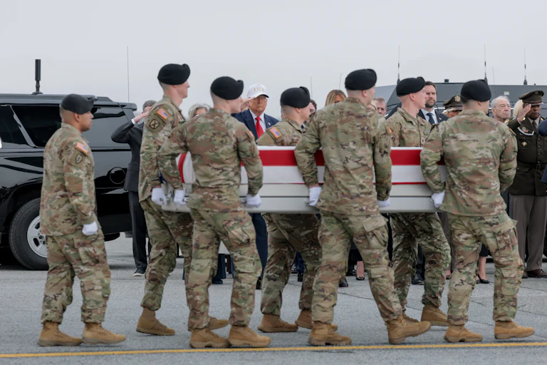 Army troops carry the American flag-covered coffin of a soldier killed in the strike on an operations center in Kuwait. Donald Trump stands in the background and salutes