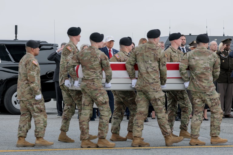 Army troops carry the American flag-covered coffin of a soldier killed in the strike on an operations center in Kuwait. Donald Trump stands in the background and salutes