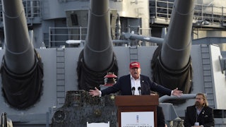 Donald Trump splays his arms outward while giving a speech aboard a battleship. He wears a red MAGA cap.