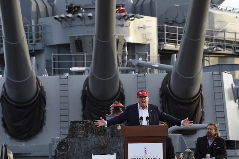 Donald Trump splays his arms outward while giving a speech aboard a battleship. He wears a red MAGA cap.