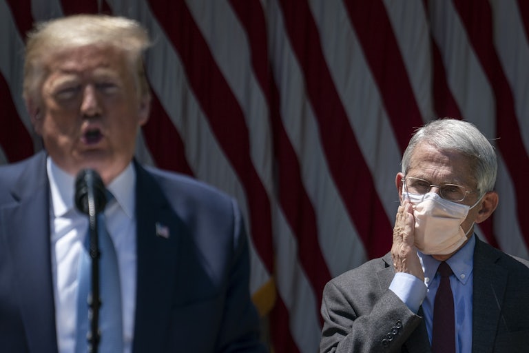 Dr. Anthony Fauci in the background wears a mask and puts a hand on his cheek, as if in exasperation or surprise. Donald Trump is in the foreground (out of focus) not wearing a mask.