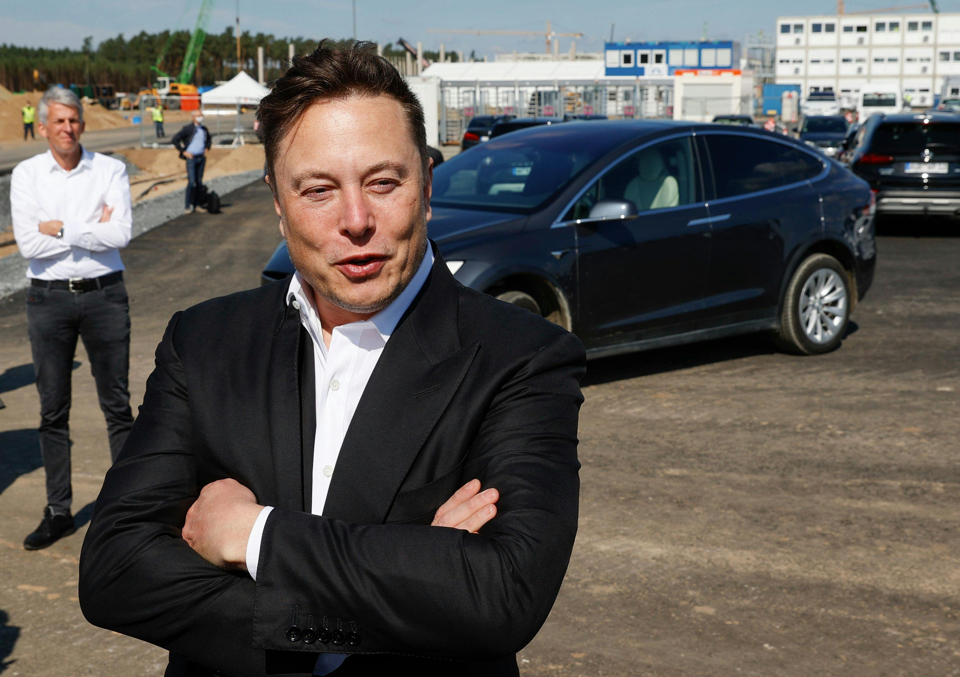 Elon Musk crosses his arms over his chest as he stands in front of a Tesla