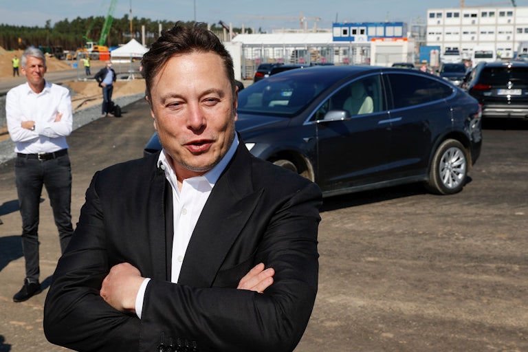 Elon Musk crosses his arms over his chest as he stands in front of a Tesla