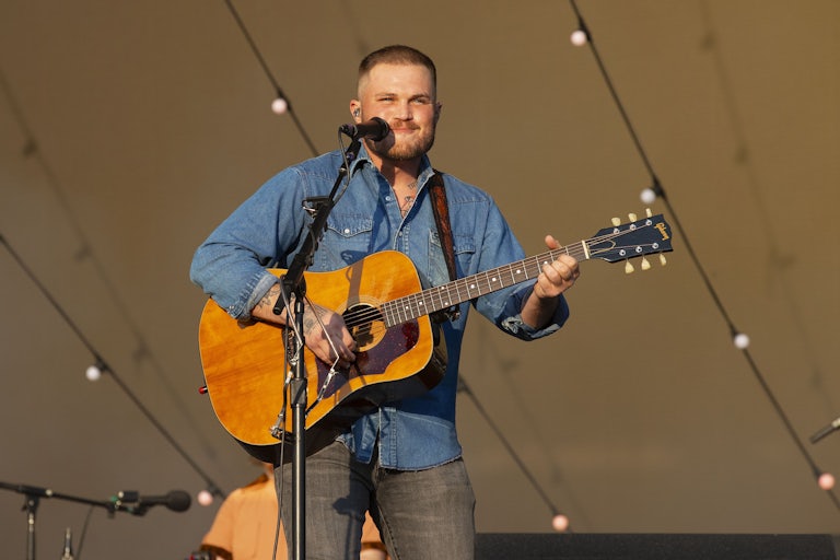 Zach Bryan plays a guitar on stage while standing at a microphone