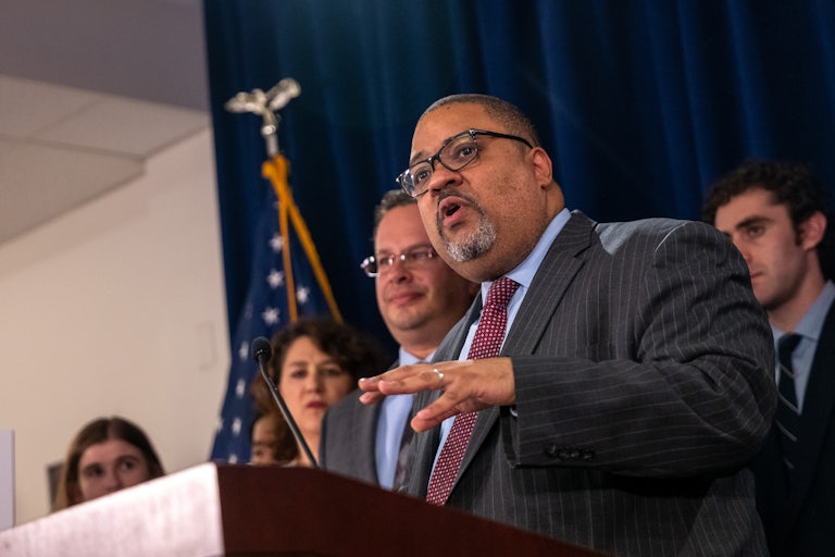 Manhattan D.A. Alvin Bragg speaks at a lectern and makes a hand gesture. Others stand by him in the background.