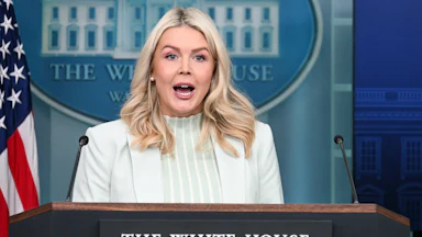 Karoline Leavitt stands behind a podium with "The White House, Washington" written on it while wearing a white blazer and shirt. Behind her is a blue placard with the White House on it.