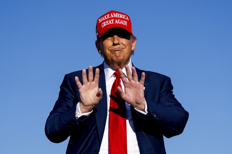 Donald Trump purses his lips and holds his palms up while wearing a read "Make America Great Again" hat. 