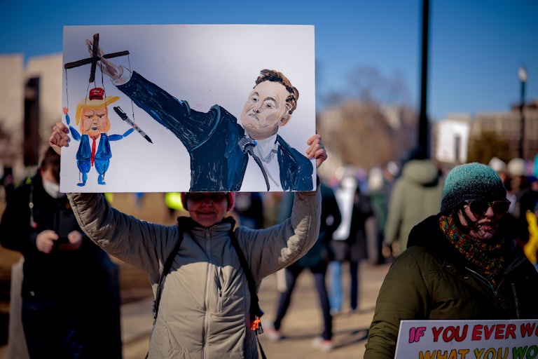 A protester holds up a drawing of Elon Musk doing a Nazi salute. His extended hand holds a puppet of Donald Trump