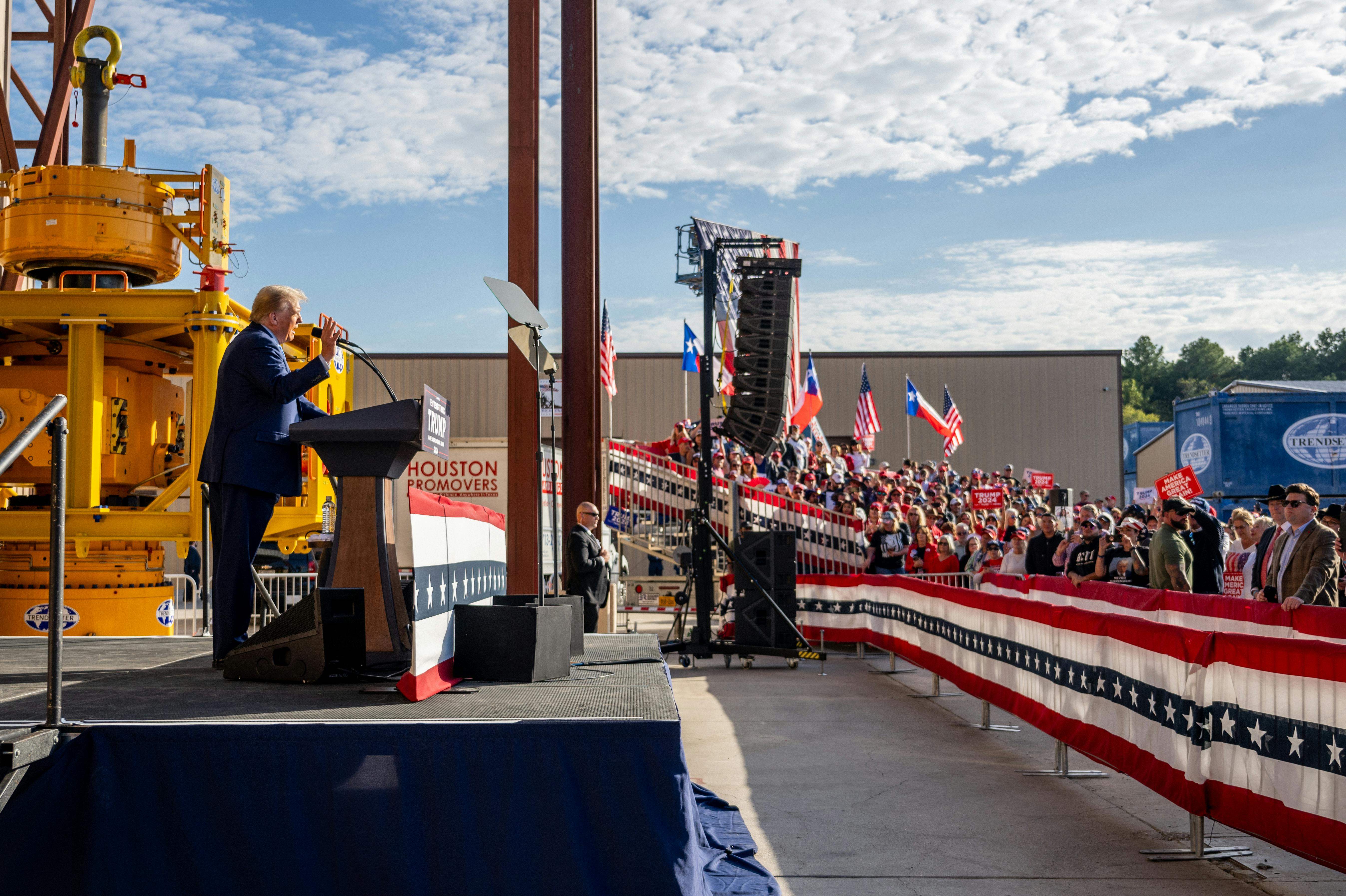 Trump stands in profile at a podium, speaking to a crowd of people.