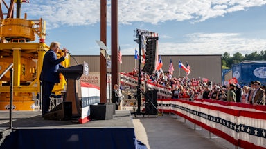 Trump stands in profile at a podium, speaking to a crowd of people.