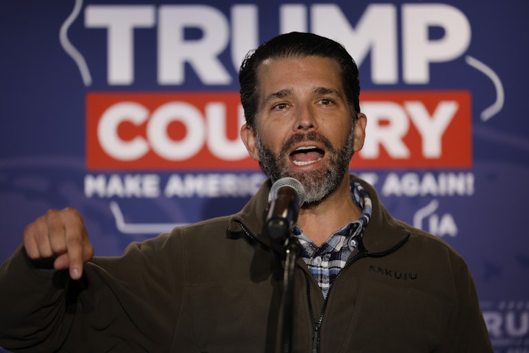 Donald Trump Jr. speaks at a mic and makes a hand gesture pointing down. A backdrop reads "Trump Country Make America Great Again!"