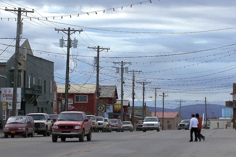 Cars drive down a road lined with small businesses, with hills in the background.