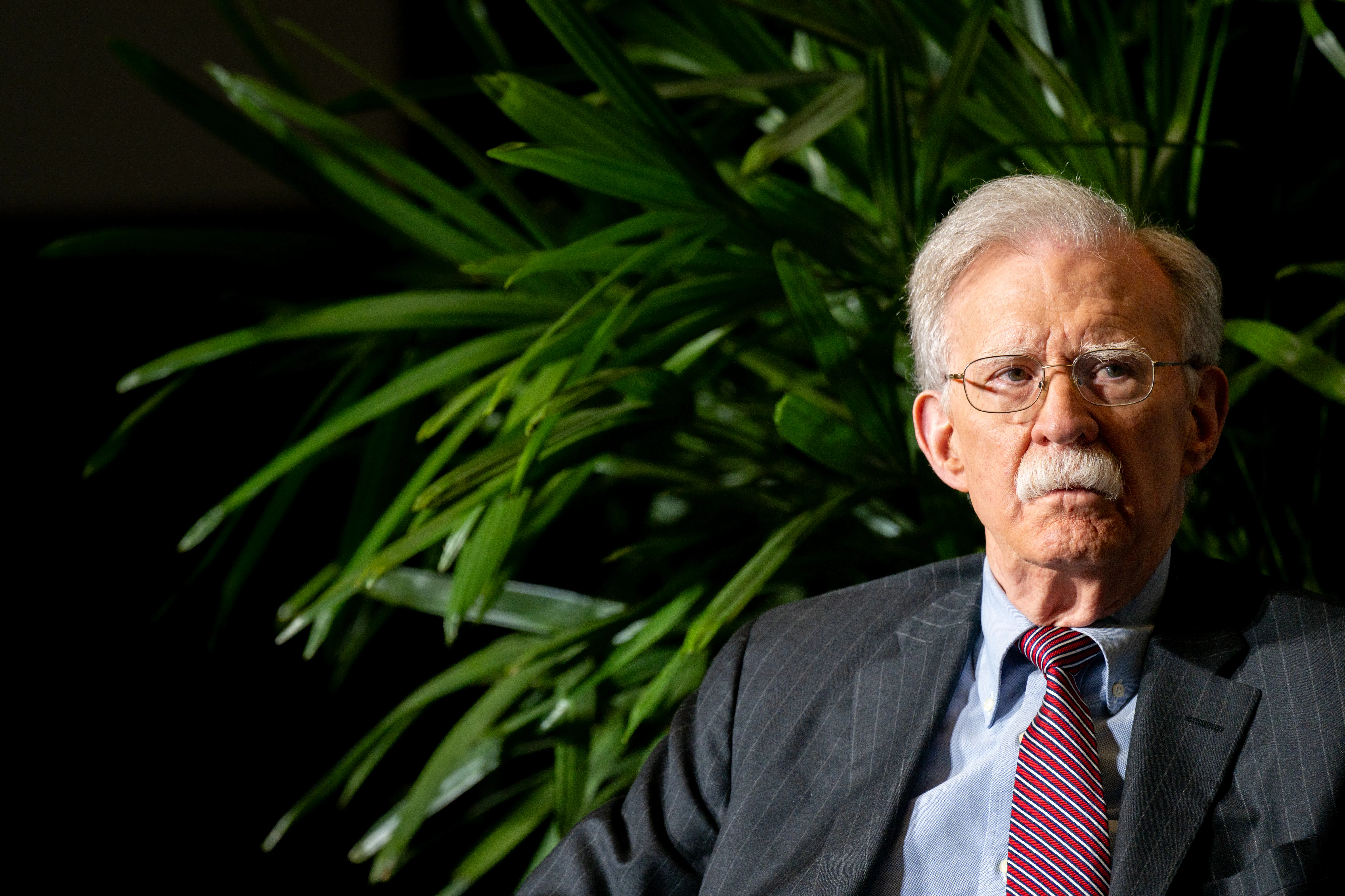 John Bolton sits in front of a plant onstage during an event