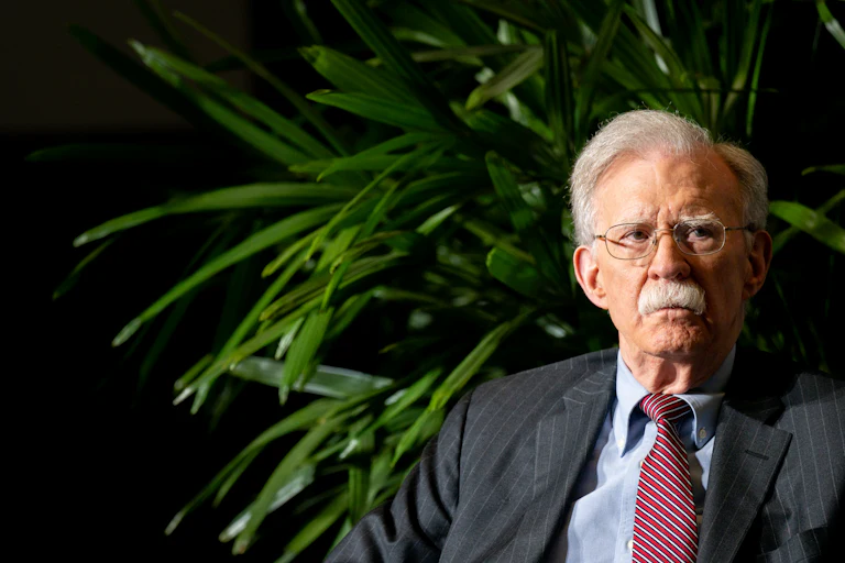 John Bolton sits in front of a plant onstage during an event