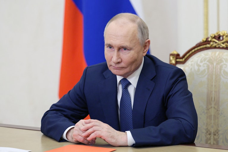 Russian leader Vladimir Putin at a table with several papers in front of him and the Russian flag in the background.