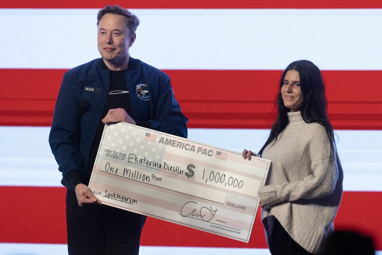 Elon Musk and Ekaterina Diestler hold a very large $1 million check made out to her. The check comes from America PAC. Both of them smile in front of a very large U.S. flag.