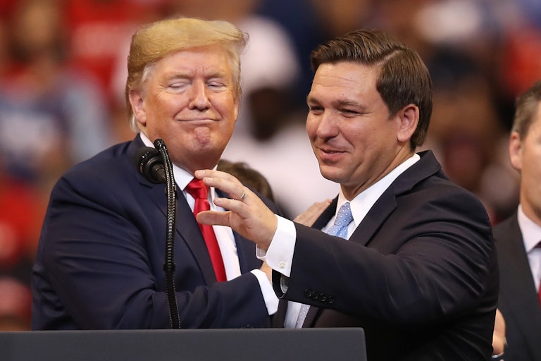 Donald Trump puts his hand on Ron DeSantis’s shoulder as he reaches for the podium mic. Both smile weirdly.