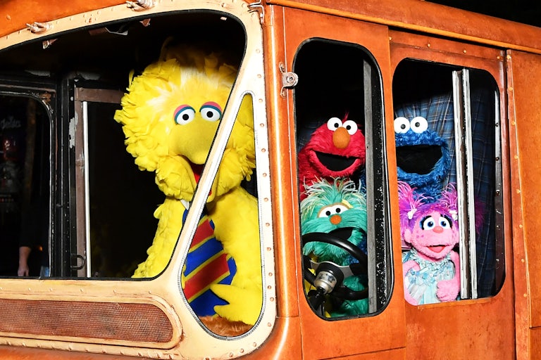 Sesame Street charactes Big Bird, Elmo, Cookie Monster, and Abby Cadabby smile in the windows of a bus. Big Bird is the bus driver.