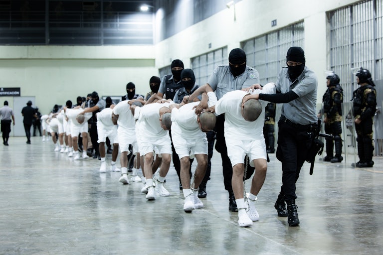 Maked agents force a row of men to bend over and walk forward, as their hands are behind their back and handcuffed to their feet.