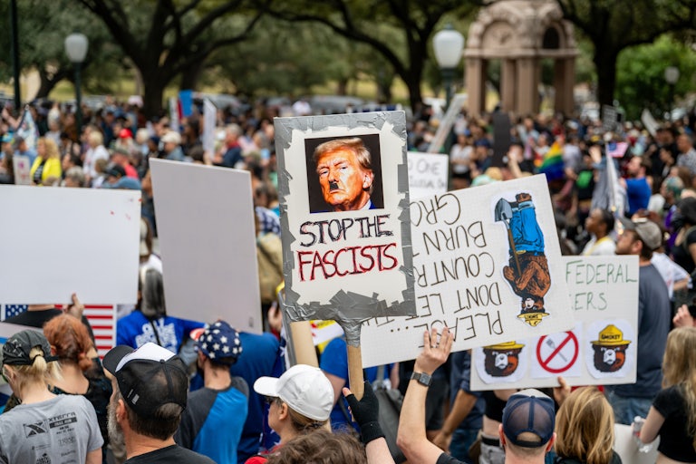 A crowd of people gather in protest. One sign reads "Stop the Fascists" and has a picture of Donald Trump with a Hitler mustache.