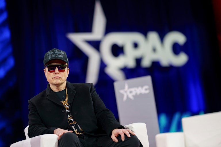 Elon Musk sitting on a chair on the stage at CPAC. He's dressed like a tool with a black MAGA hat, a heavy gold chain, and absurd red and black sunglasses.