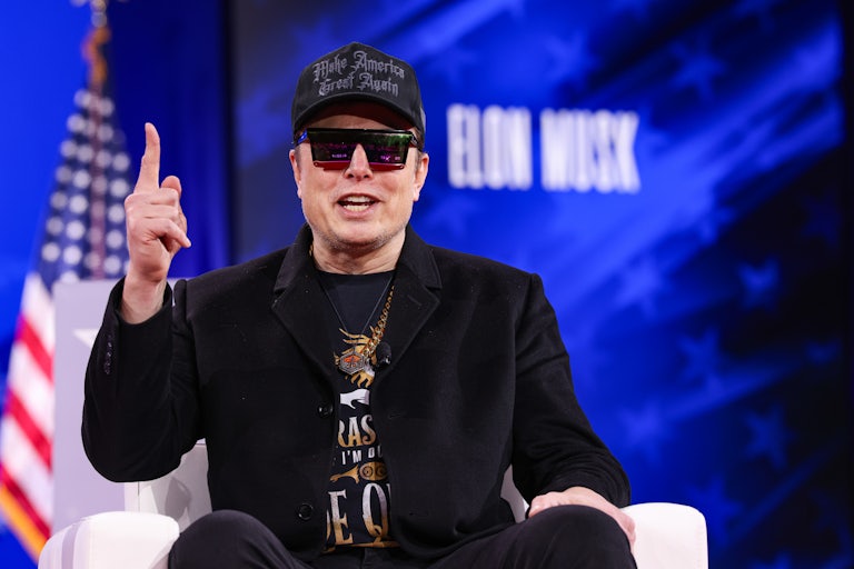 Elon Musk speaks at CPAC. He's dressed ridiculously, with black and red sunglasses (the event is indoors), a black MAGA cap, a heavy gold chain, andn a black jacket over a graphic tee.