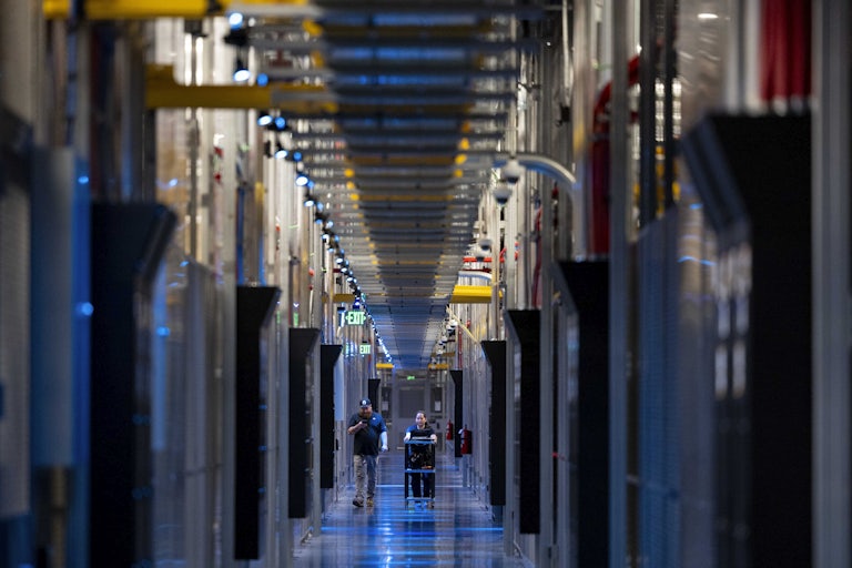 Two people walk down a corridor with columns of electronic equipment on either side. One pushes a cart.