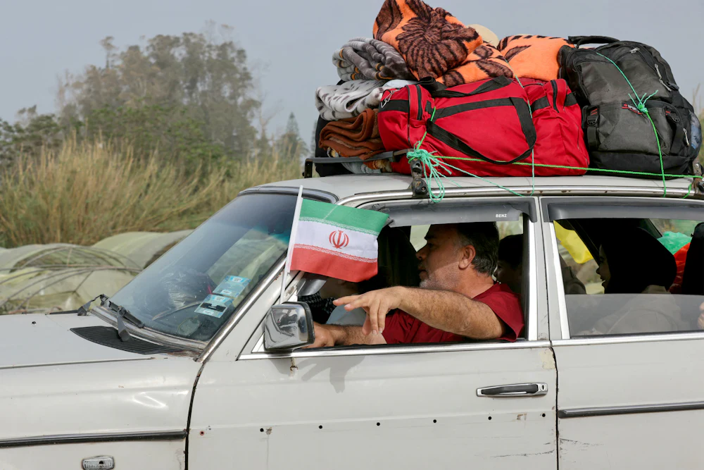 A man holds an Iranian flag as displaced residents make their way back to their homes on a makeshift road, built at the site where the Qasmieh bridge was destroyed in Israeli strikes, in the southern Lebanese area of Al-Qasmiyeh on April 18, 2026.