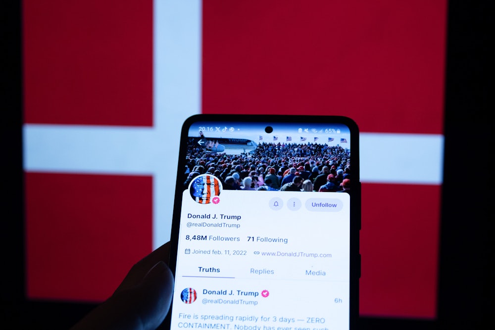 In this photo illustration, President-elect Donald Trump's account on Truth Social is seen displayed on a smartphone, and the flag of Denmark is seen on a laptop screen in the background.