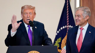 Donald Trump raises one hand while speaking at a podium. Dr. Oz stands to his left and smiles at him.