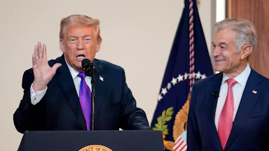 Donald Trump raises one hand while speaking at a podium. Dr. Oz stands to his left and smiles at him.