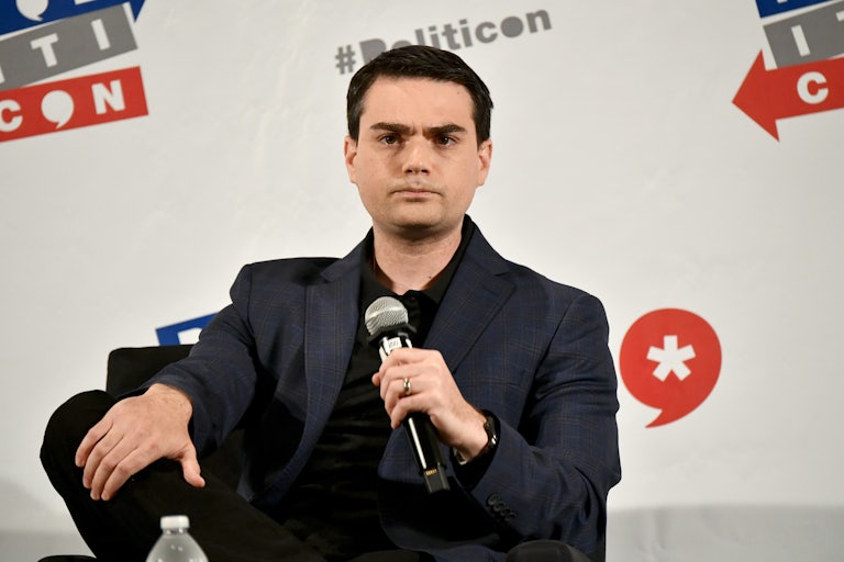 Ben Shapiro rests his hand on his leg and sits in front of a Politicon backdrop