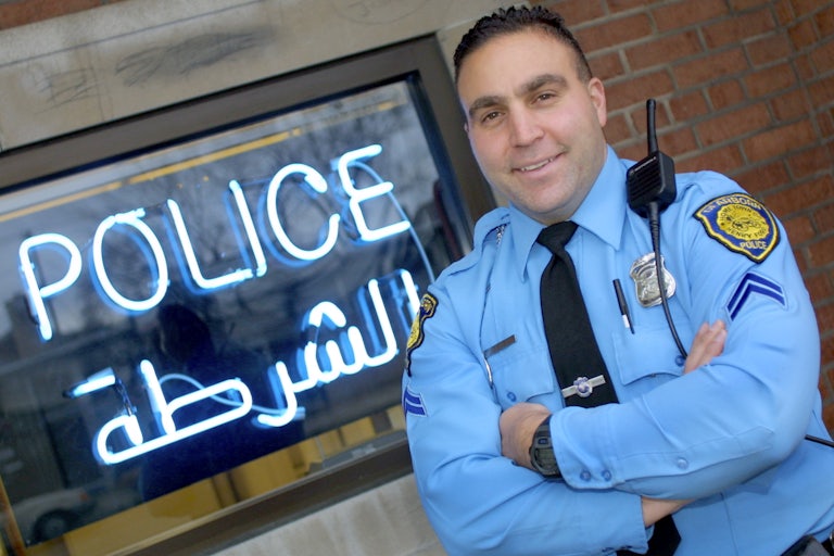 A Dearborn police officer in uniform stands in front of a sign that reads Police in Arabic ad English.