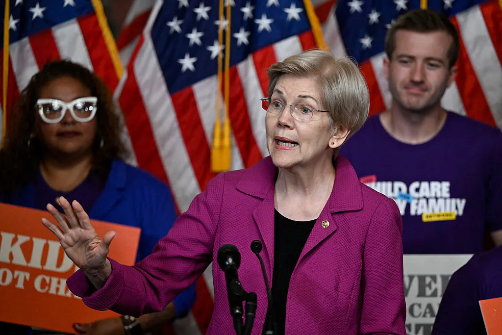 Elizabeth Warren gestures as people with signs stand behind her.