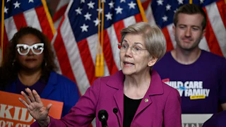 Elizabeth Warren gestures as people with signs stand behind her.