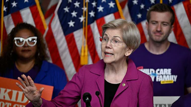 Elizabeth Warren gestures as people with signs stand behind her.