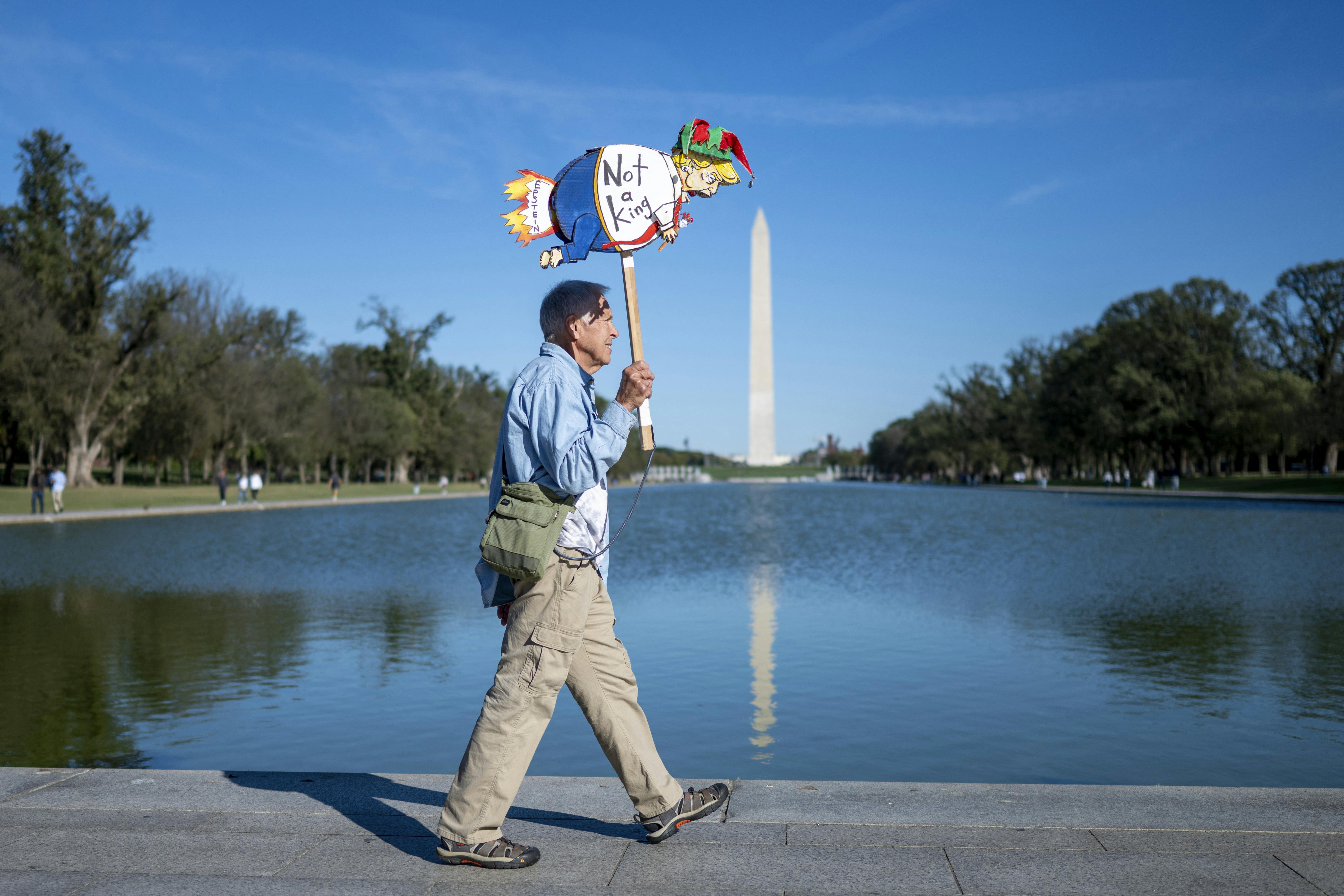 A protester walks past the Reflecting Pool, opposite the Washington Monument, holding up a drawing of Donald Trump that says, "Not a king"