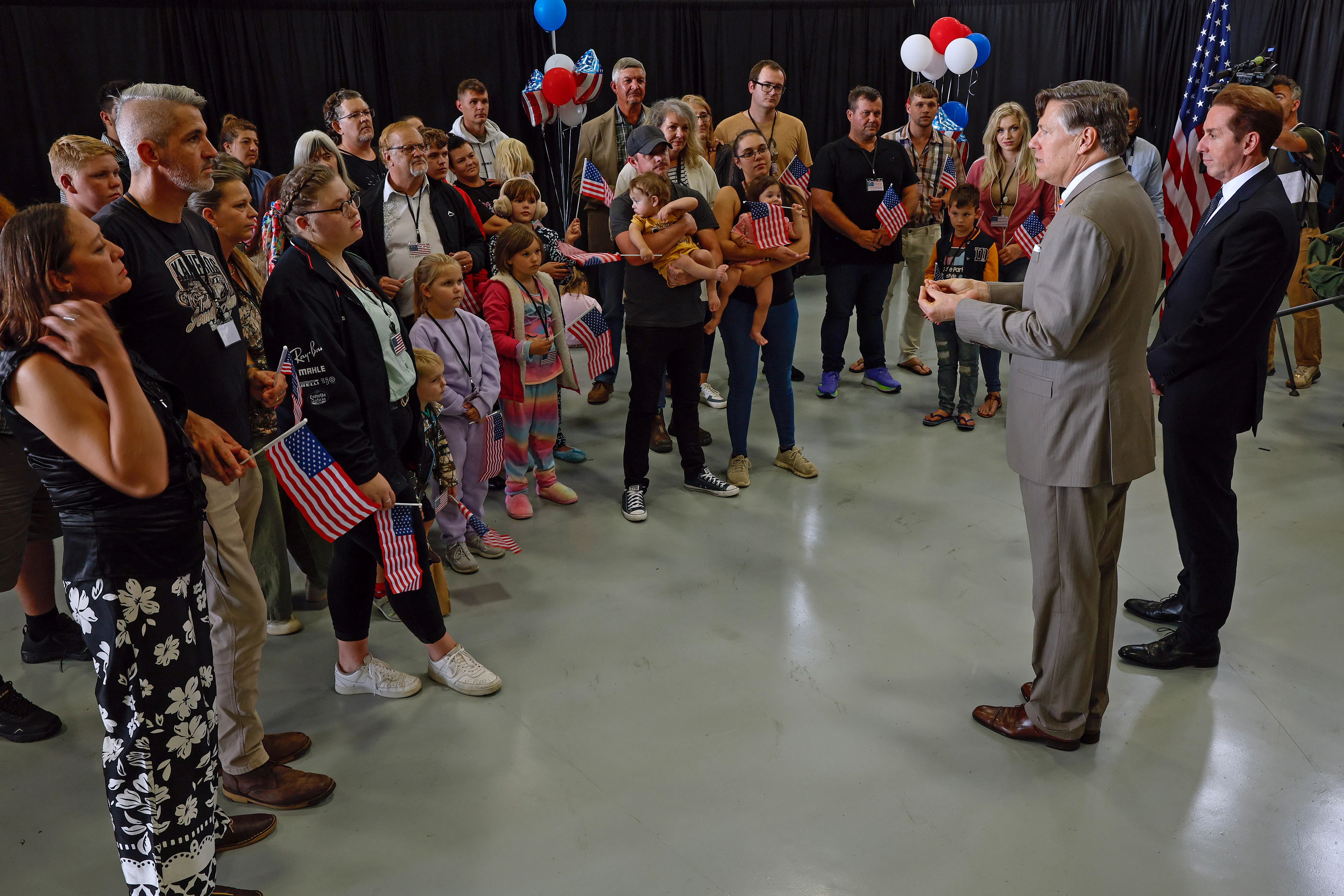 A crowd of white South Africans holding American flags and balloons listen to two men wearing suits in an airplane hangar. 