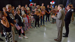 A crowd of white South Africans holding American flags and balloons listen to two men wearing suits in an airplane hangar.