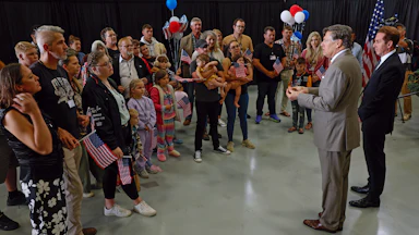 A crowd of white South Africans holding American flags and balloons listen to two men wearing suits in an airplane hangar.