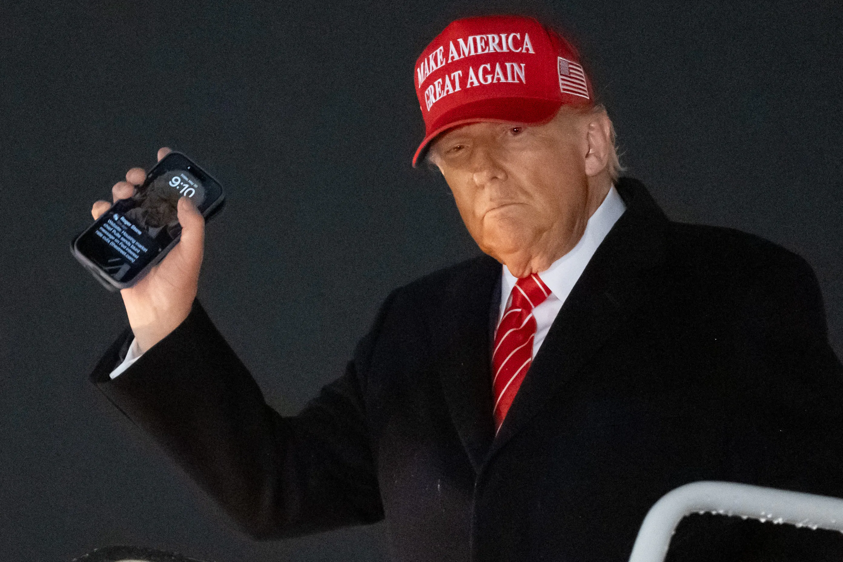 Donald Trump wears a red Make America Great Again hat and holds up his phone.