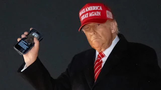 Donald Trump wears a red Make America Great Again hat and holds up his phone.