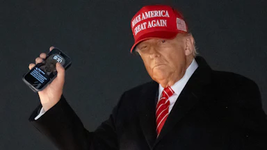 Donald Trump wears a red Make America Great Again hat and holds up his phone.
