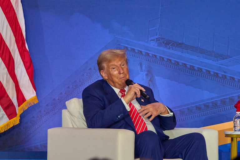 Donald Trump sits in an armchair and holds a microphone to his face during a Moms for Liberty event