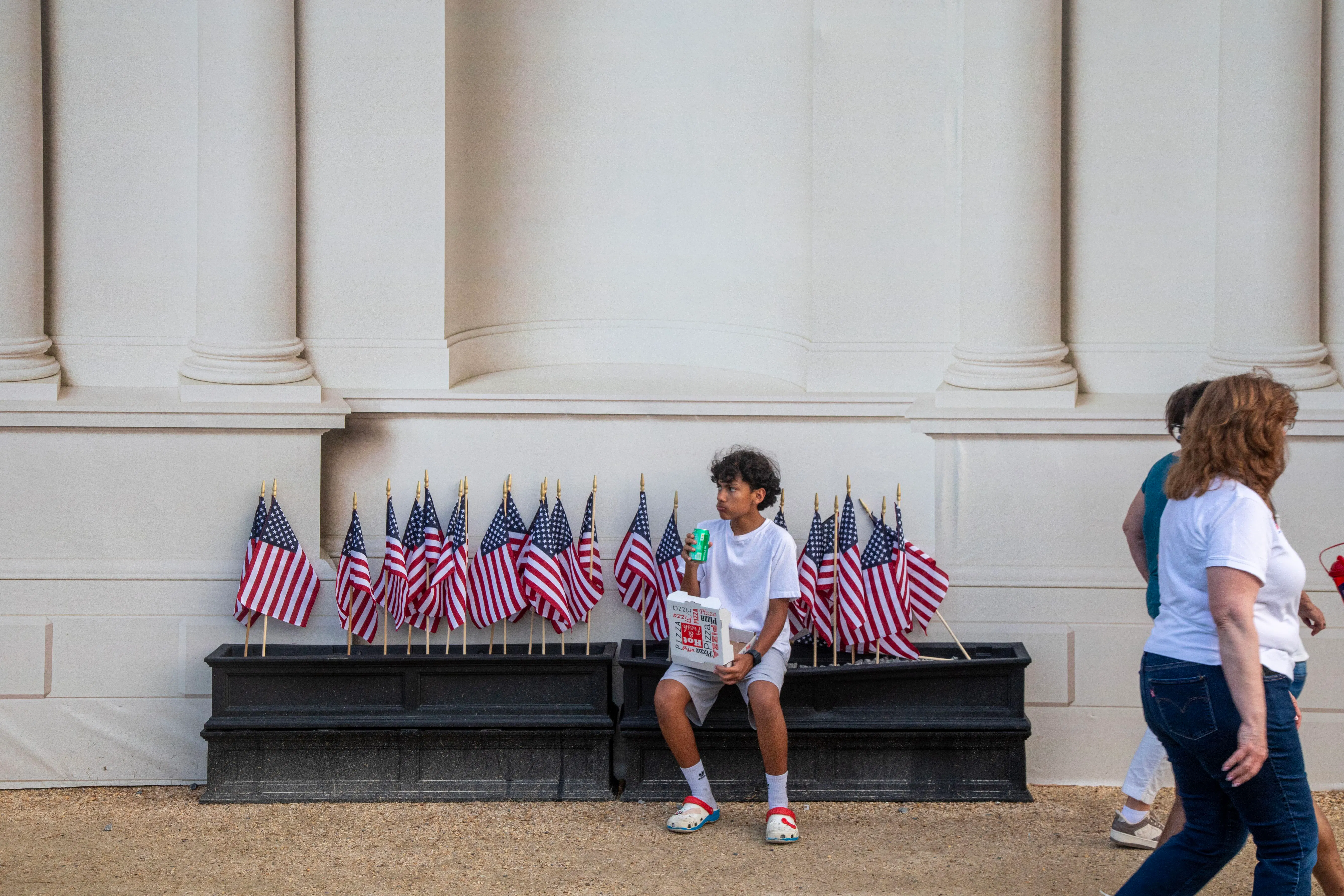 A boy sits on black boxes carrying small American flags at the base of a building with white columns. The boy is wearing a white t-shirt and light-colored shorts. 