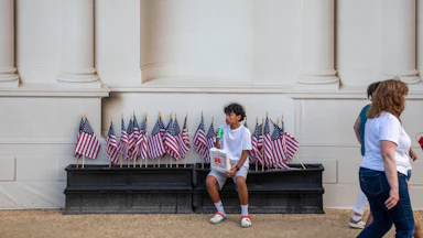 A boy sits on black boxes carrying small American flags at the base of a building with white columns. The boy is wearing a white t-shirt and light-colored shorts.